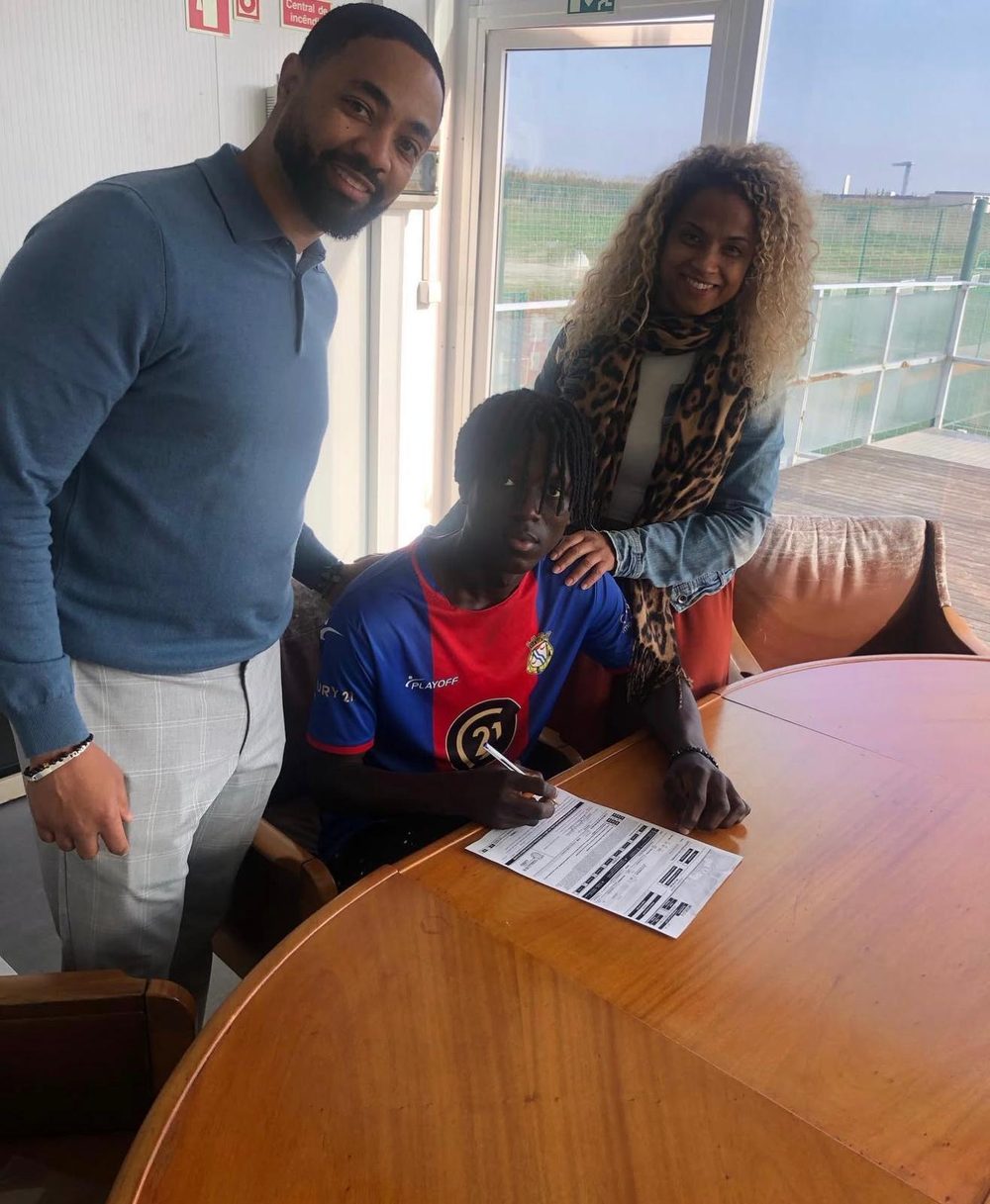 A young player in an FC Alverca shirt signing a document at a table, with a man and a woman standing beside him