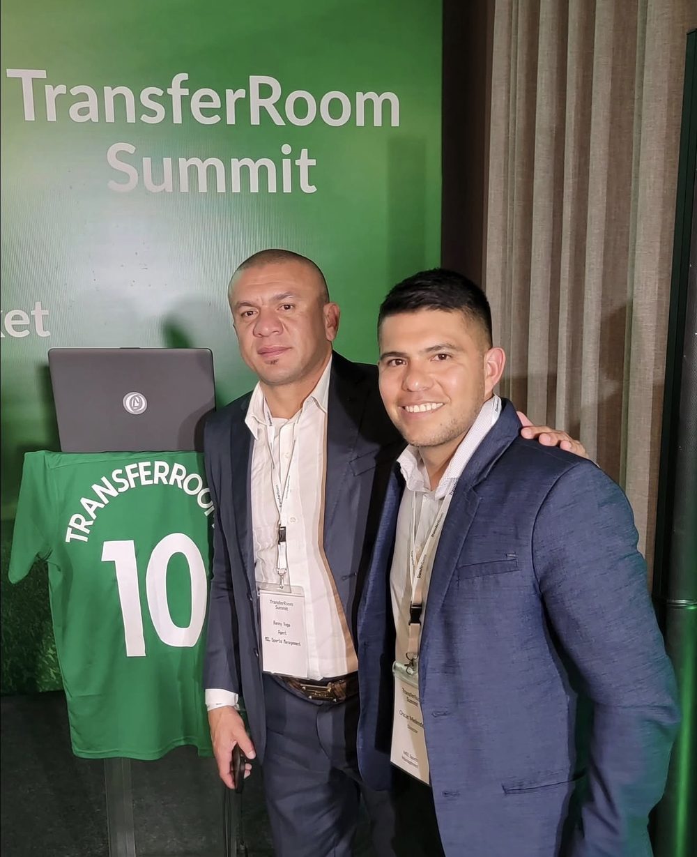 Two men in suits with event badges in front of a TransferRoom Summit backdrop in São Paulo